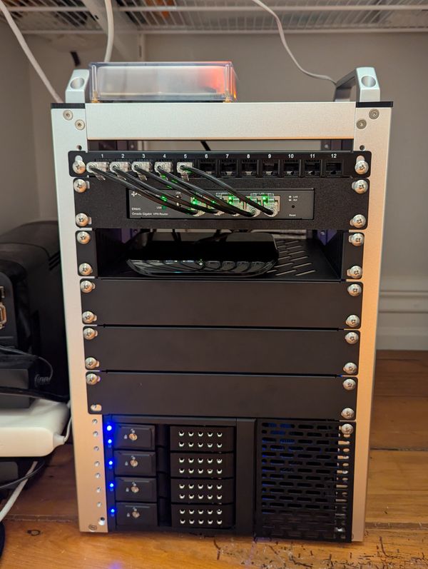 A 10-inch, silver and black rack-mounted server unit on a wooden floor. The top row has a Home Assistant Yellow with a glowing light, below it a patch panel with Ethernet cables connected to a router showing green status lights. The lower sections house several blank panels, drive bays with lockable trays, and a vented cooling panel.