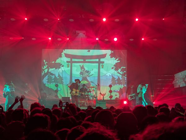 A band performs on stage under bright red lighting, with a large illustrated backdrop depicting a Japanese torii gate, mountains, and trees. The audience is silhouetted in the foreground.