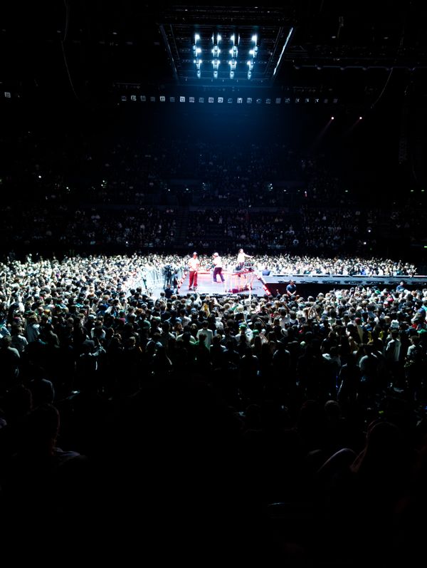 A packed arena with a brightly lit center stage featuring three performers: Paris Texas. The surrounding crowd is dense, and overhead lights illuminate the scene dramatically.
